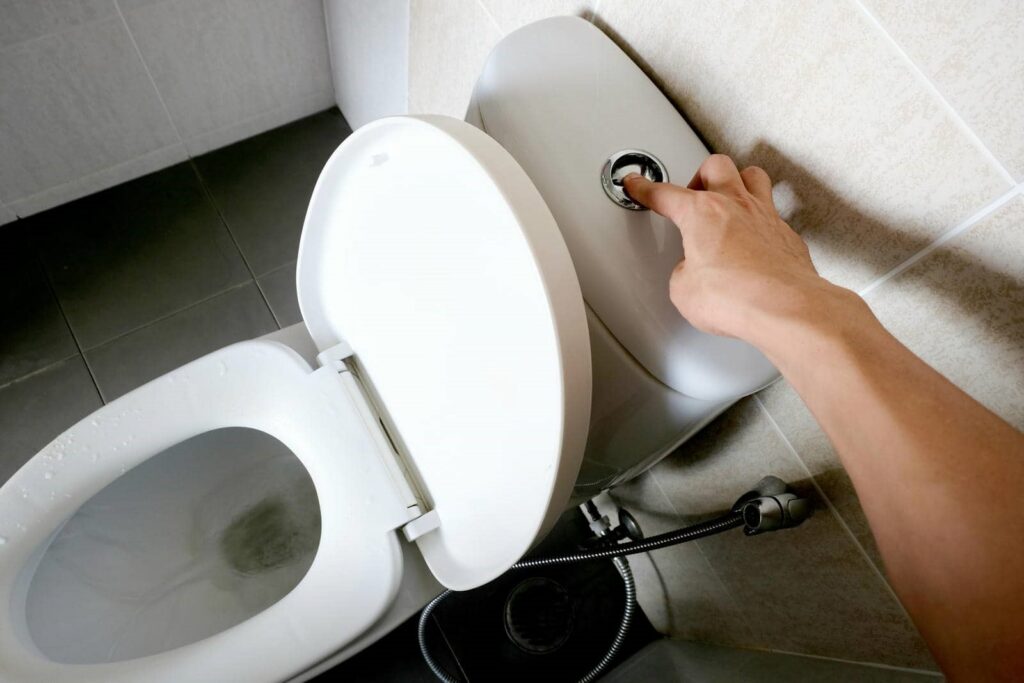 Hand pressing flush button on a white toilet with the lid open.