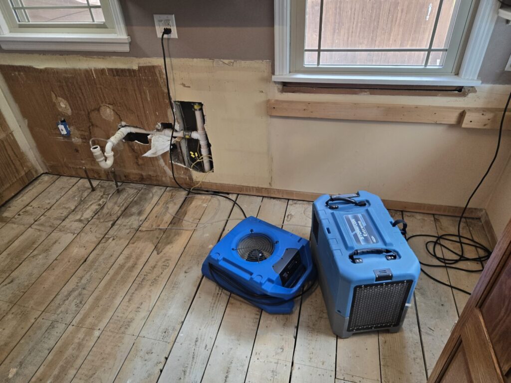 Two blue air movers or drying machines on a wooden floor in a partially stripped room.