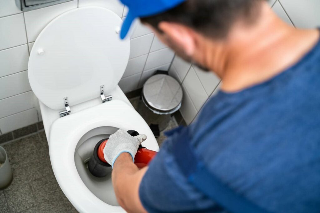 Person using a plunger to unclog a white toilet in a tiled bathroom.